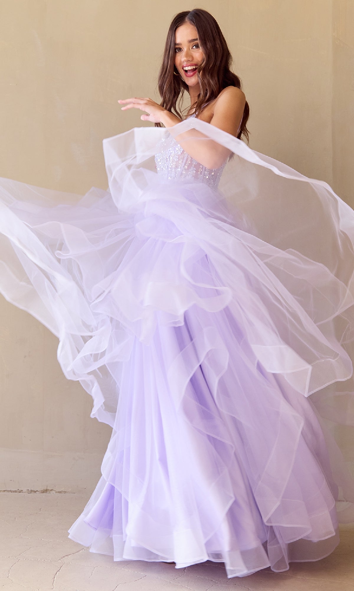A woman twirls in the Long Glitter Prom Ball Gown: Amelia Couture 5068, her light purple a-line dress sparkling as its layers flare out and she smiles against a plain beige wall.